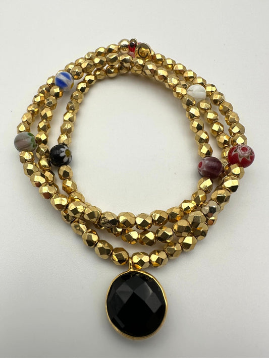 Ce bijou CaJouX est composé de facettes de Bohème couleur Or plaqué 24K sur un fil élastique de grande qualité. On y retrouve aussi des perles rondes Millefiori et ça breloque Onyx noir et plaqué Or.